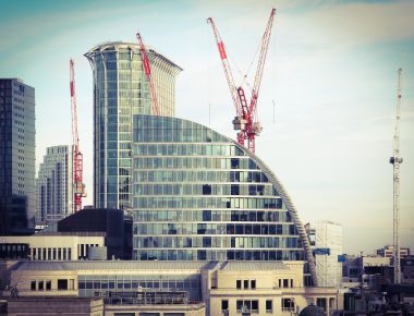 Modern skyline with glass buildings and red construction cranes against a pale sky, creating an urban scene.